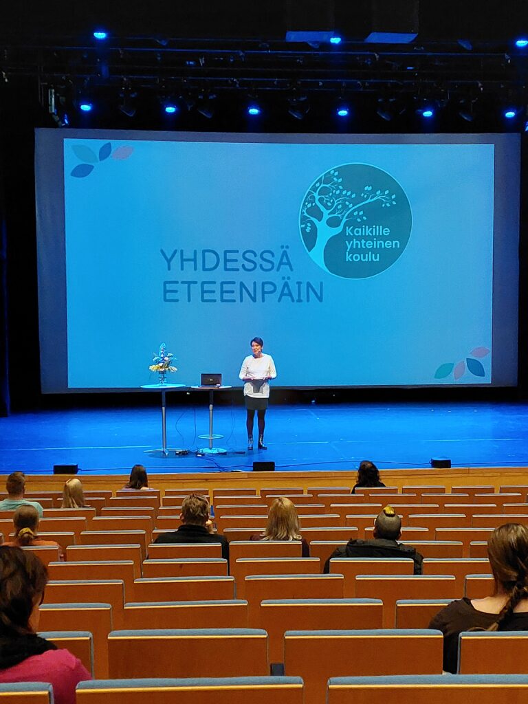 Henkilö seisoo lavalla ja katsoo yleisöön, takana esityksessä teksti "Yhdessä eteenpäin".