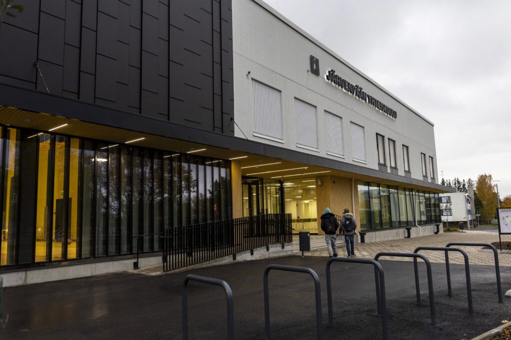 Koulurakennuksen sisäänkäynti, jossa kaksi henkilöä kävelee kohti sisäänkäyntiä. Rakennuksessa on moderni arkkitehtuuri, lasiseiniä ja mustia sekä valkoisia elementtejä.