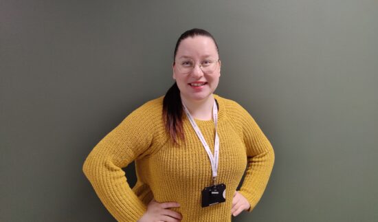 Hymyilevä henkilö seisoo vihreää seinää vasten. Hänellä on päällään sinapinkeltainen neule, silmälasit ja kaulassa henkilökortti. Kädet ovat lanteilla ja hiukset on sidottu taakse.